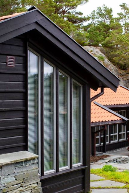 Mørkt hus med store vindu, grunnmur av sten og og med skog i bakgrunnen