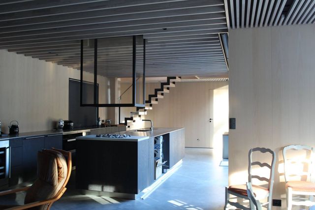 Moderne sort kjøkken i et nybygg hvor der er kjøkkentøy, en moderne enkel trapp som leder opp til andre etasje og diverse stoler i forgrunnen
